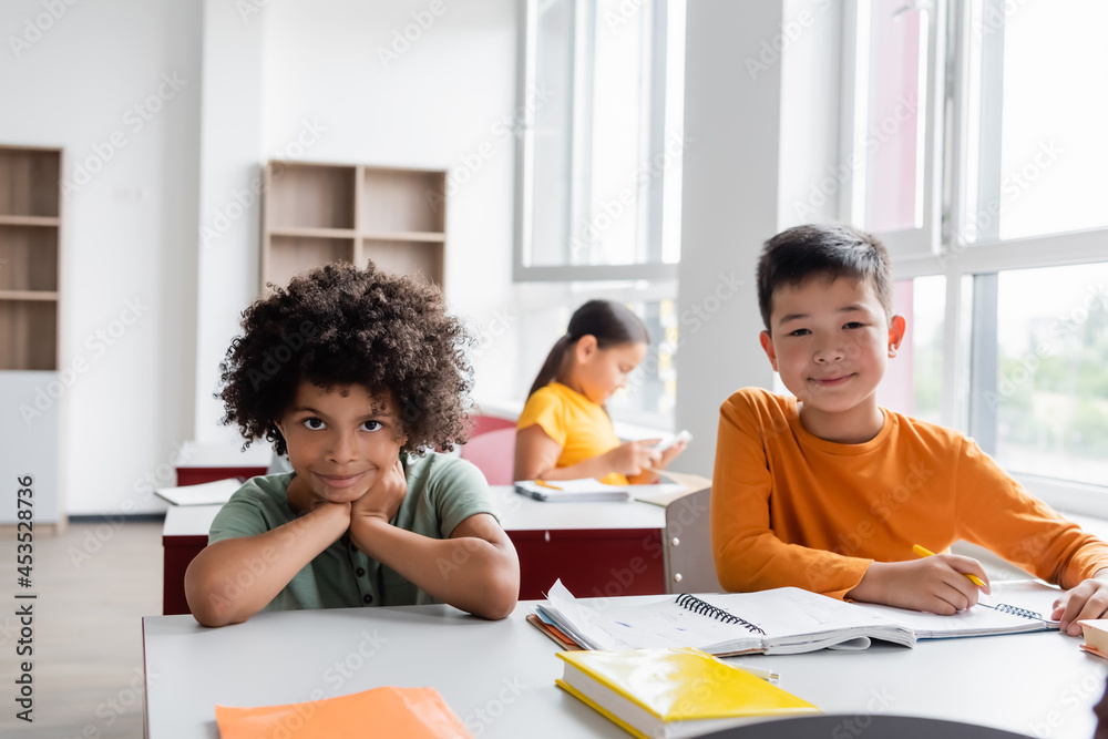 Fototapeta premium multiethnic classmates looking at camera near notebooks in classroom