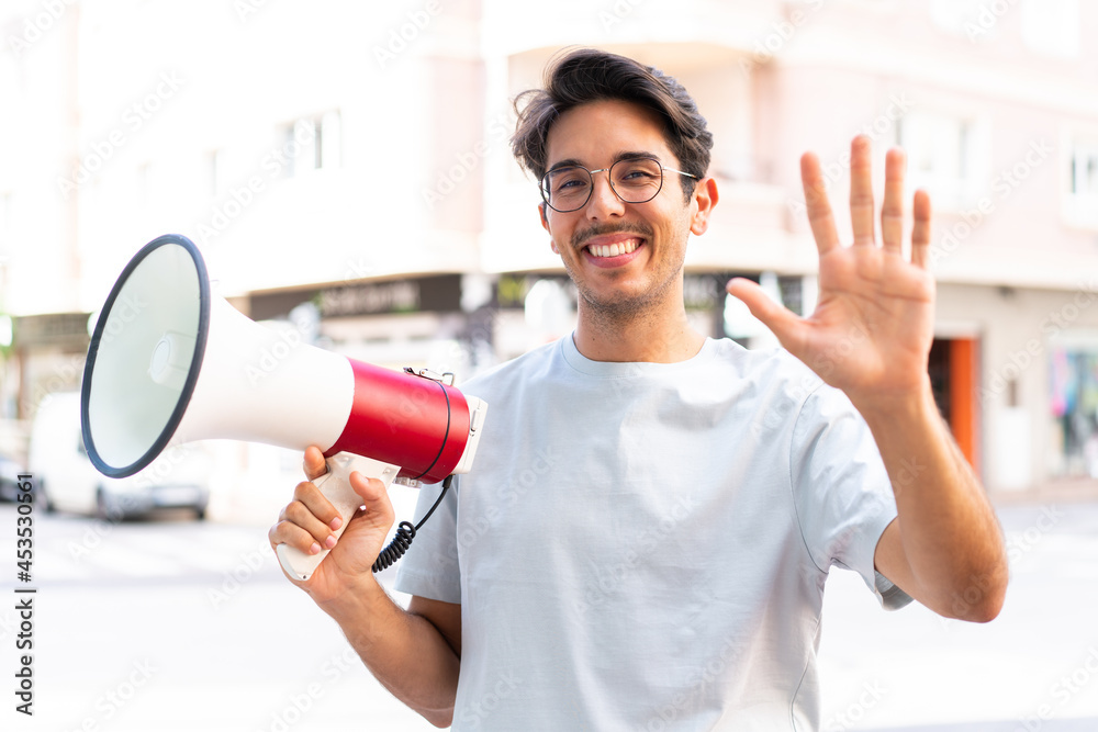 Young caucasian man at outdoors holding a megaphone and saluting with hand with happy expression Young caucasian man at outdoors holding a megaphone and saluting with hand with happy expression