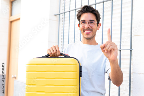Young caucasian man at outdoors in vacation with travel suitcase and counting one