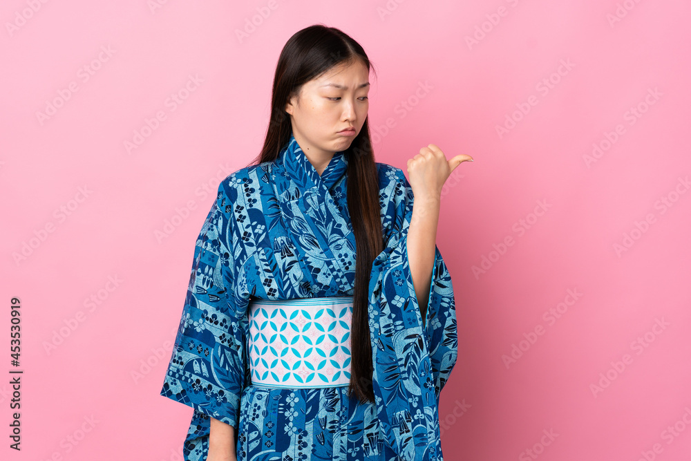 Young Chinese girl wearing kimono over isolated background unhappy and pointing to the side Young Chinese girl wearing kimono over isolated background unhappy and pointing to the side