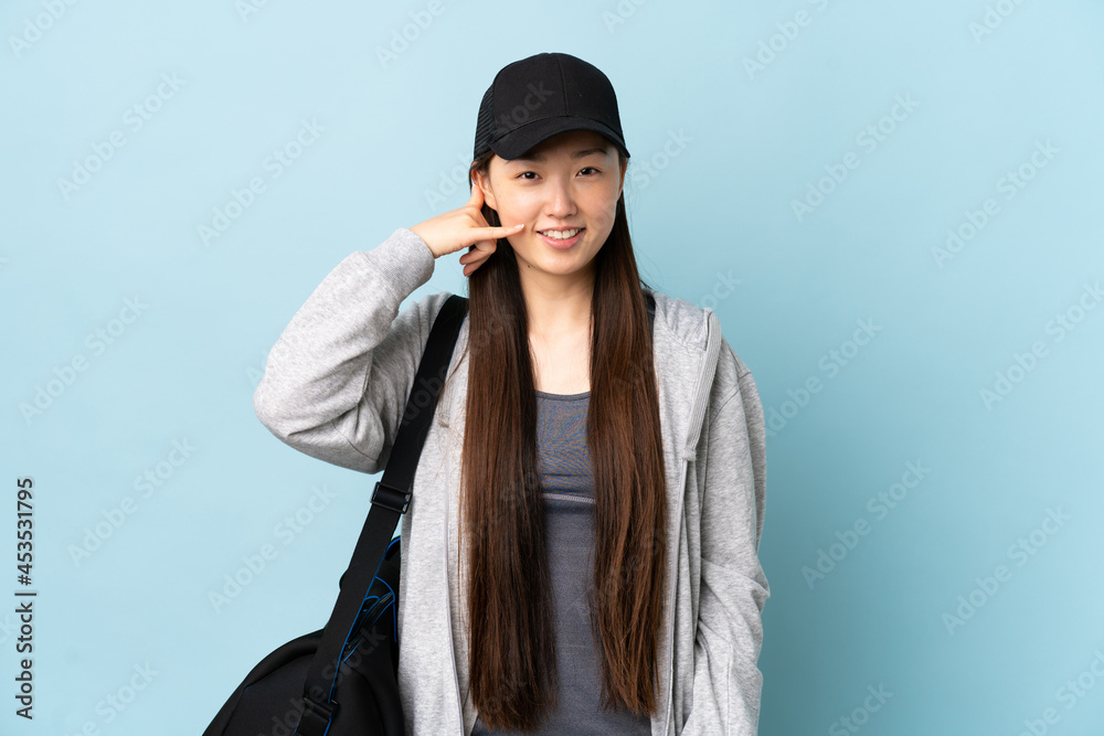 Young sport Chinese woman with sport bag over isolated blue background making phone gesture. Call me back sign Young sport Chinese woman with sport bag over isolated blue background making phone gesture. Call me back sign