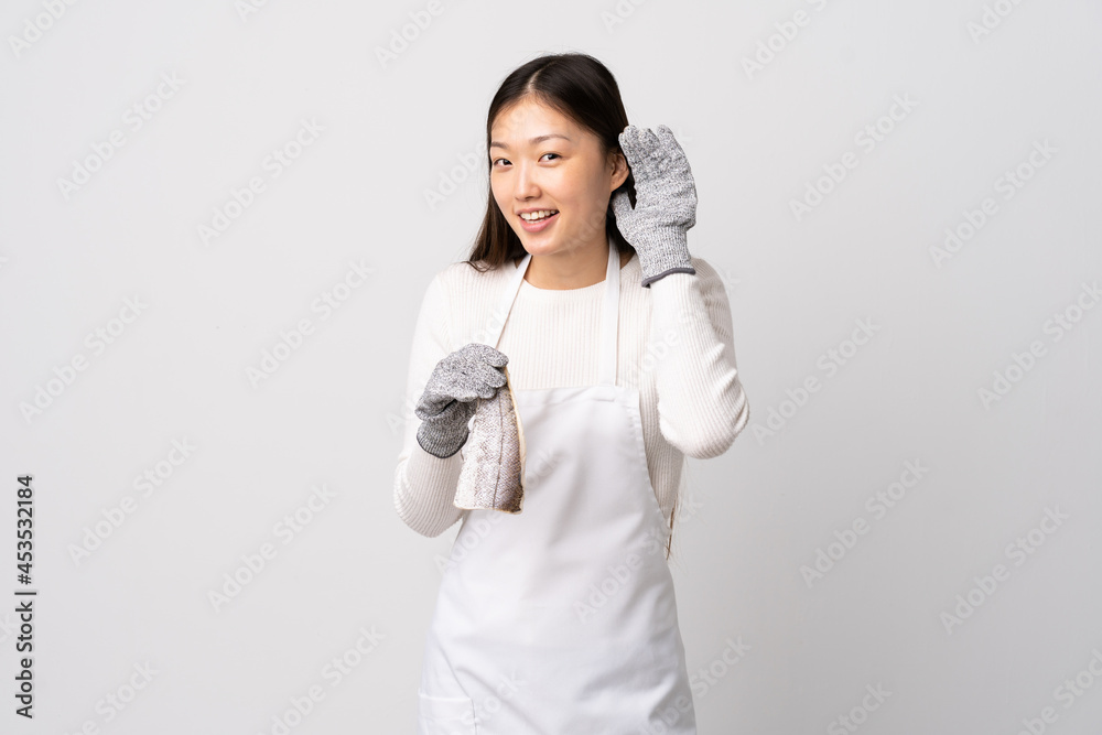 Chinese fishmonger wearing an apron and holding a raw fish over isolated white background listening to something by putting hand on the ear Chinese fishmonger wearing an apron and holding a raw fish over isolated white background listening to something by putting hand on the ear