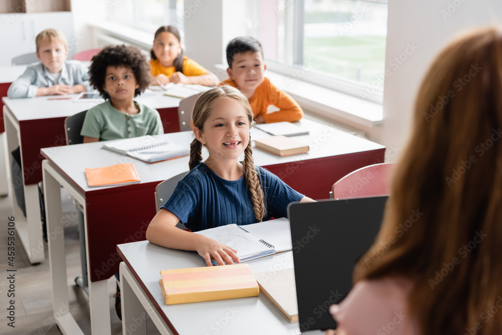 cheerful interracial kids looking at schoolgirl with laptop on blurred foreground cheerful interracial kids looking at schoolgirl with laptop on blurred foreground