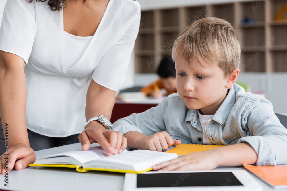 african american teacher pointing with finger at book near schoolboy and digital tablet african american teacher pointing with finger at book near schoolboy and digital tablet