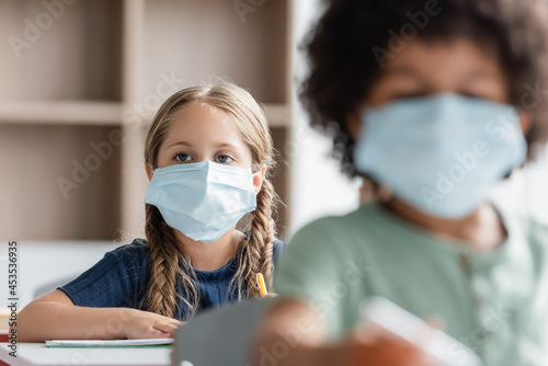 schoolgirl in medical mask near blurred african american boy in classroom
