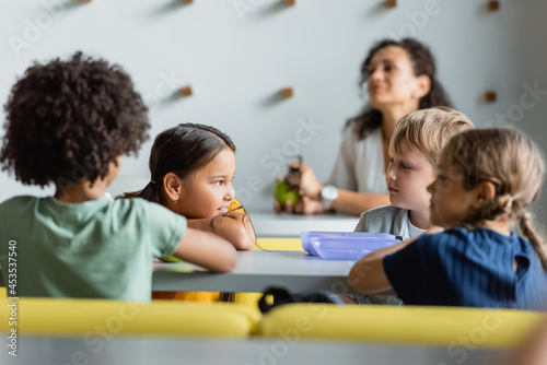 asian girl talking to friend near blurred multiethnic classmates in school eatery
