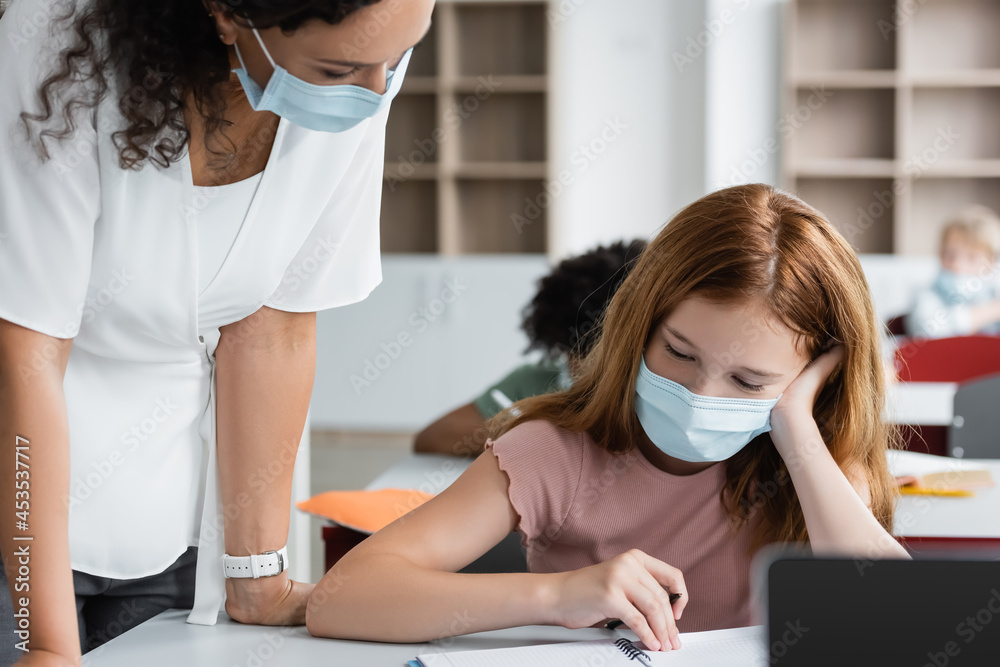 african american teacher in medical mask standing near girl looking at notebook during lesson african american teacher in medical mask standing near girl looking at notebook during lesson