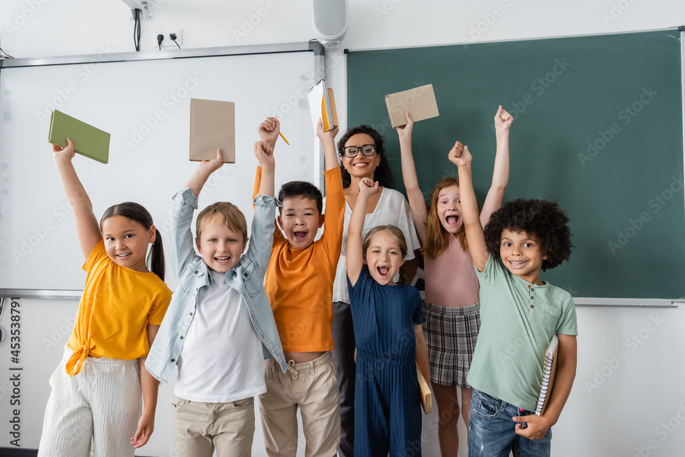 Fototapeta premium excited multiethnic schoolkids holding books in raised hands near happy african american teacher
