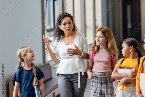 african american teacher gesturing while speaking to multicultural children in school corridor