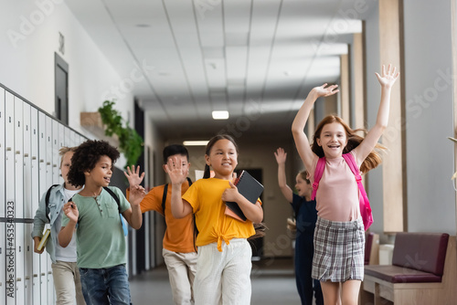 happy multiethnic pupils waving hands while running in school corridor