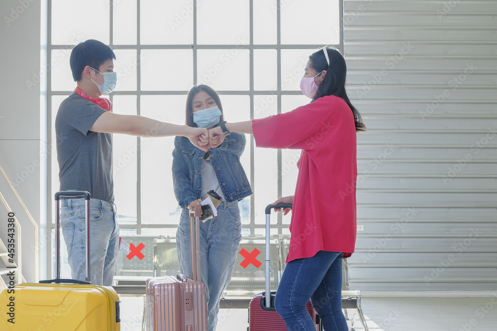 Asian young traveler group wearing protective face mask and fist bump greeting together at public terminal area. COVID-19 awareness and new normal travel lifestyle concept. Selective focus.