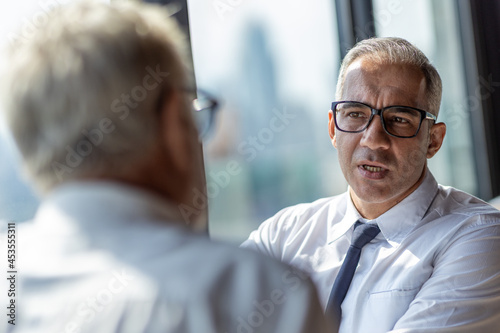 Picture of young business man taking to his older business partner. They are in white shirt and black tie. They are in a hotel lobby. 