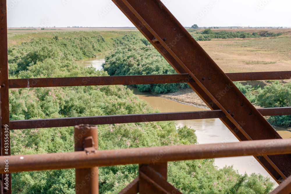the river flowing between the banks overgrown with branches, the view through the iron pillars of the bridge the river flowing between the banks overgrown with branches, the view through the iron pillars of the bridge