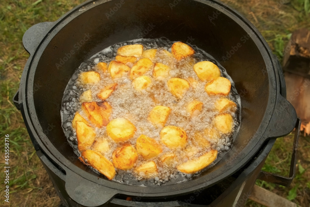 Potato is frying in cauldron in oil on nature on fire, closeup view. Preparing food on campfire in camping. Large pieces of potatoes are deep-fried in a black kazan. Cooking using wood-burning stove. Potato is frying in cauldron in oil on nature on fire, closeup view. Preparing food on campfire in camping. Large pieces of potatoes are deep-fried in a black kazan. Cooking using wood-burning stove.