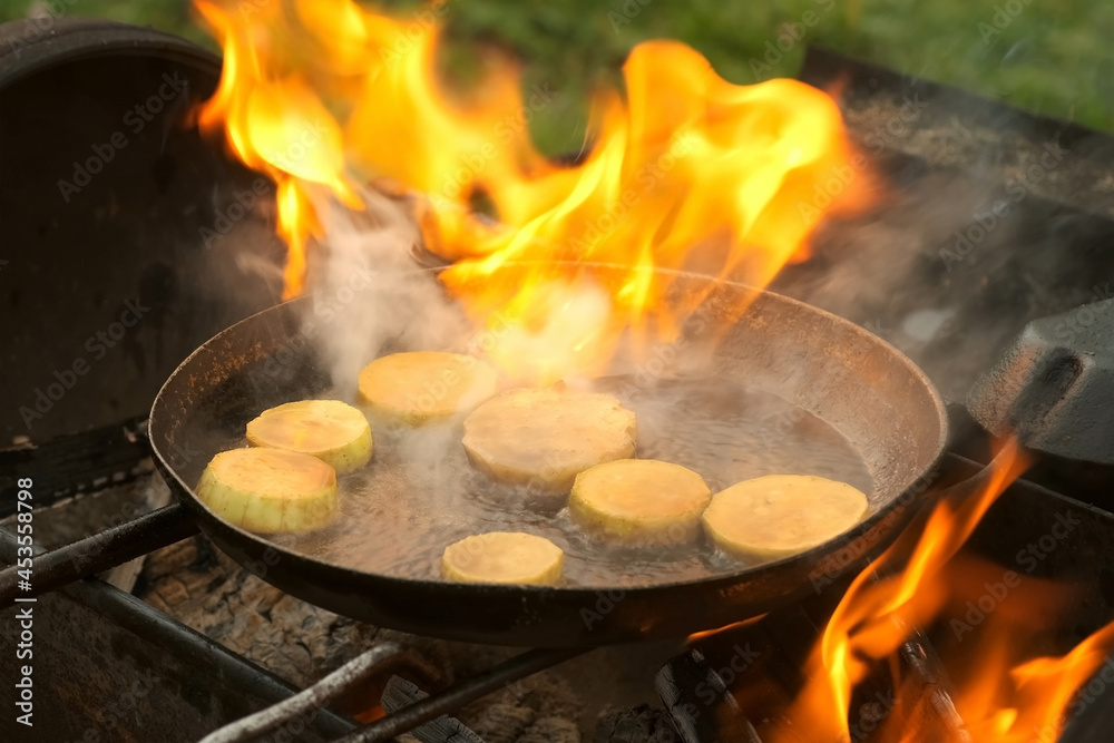 Man is frying slices of fresh organic zucchini on frying pan with hot boiling oil on coals in grill. He is putting zucchini in hot oil and its starting to burn. Preparing food in chargrill on nature. Man is frying slices of fresh organic zucchini on frying pan with hot boiling oil on coals in grill. He is putting zucchini in hot oil and its starting to burn. Preparing food in chargrill on nature.