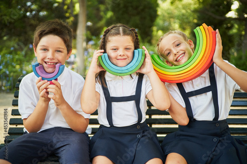 Three children wearing school uniform smiling at camera with rainbow toy in park