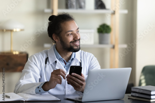 Dreamy happy young african american mixed race gp doctor therapist physician holding cellphone in hands, sitting at table with computer in modern clinic office room, resting during hard workday.