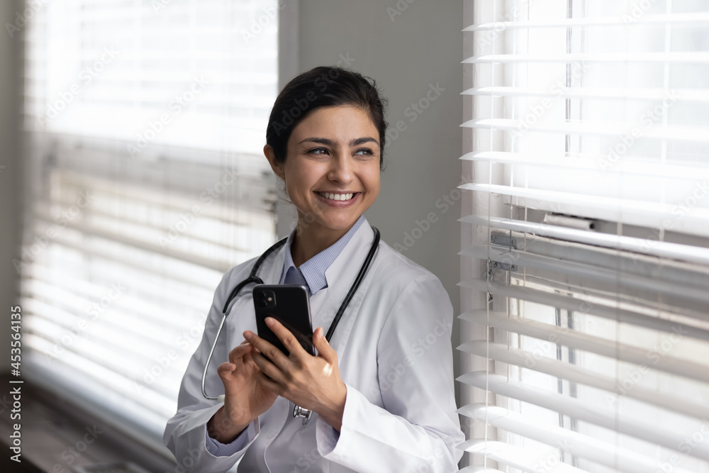 Smiling dreamy beautiful young indian ethnicity female gp doctor holding cellphone in hands, standing near window in modern clinic office, resting during workday or thinking on getting good news. Smiling dreamy beautiful young indian ethnicity female gp doctor holding cellphone in hands, standing near window in modern clinic office, resting during workday or thinking on getting good news.