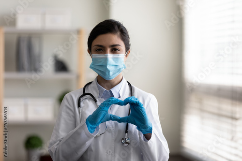 Happy young indian ethnicity female gp doctor infectious disease specialist in disposable gloves and protective facial mask making heart sign, expressing support to ill patient, charity concept. photo