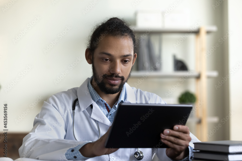 Interested concentrated young african american male gp doctor physician holding digital tablet, web surfing useful information, consulting patient distantly or using medical healthcare application. Interested concentrated young african american male gp doctor physician holding digital tablet, web surfing useful information, consulting patient distantly or using medical healthcare application.