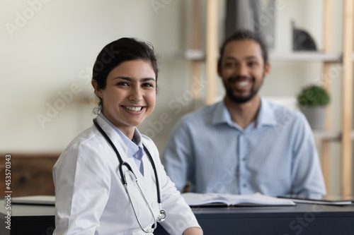 Portrait of smiling confident young female indian ethnicity gp doctor physician posing in clinic with blurred african american male patient on background, professional medicare healthcare services.