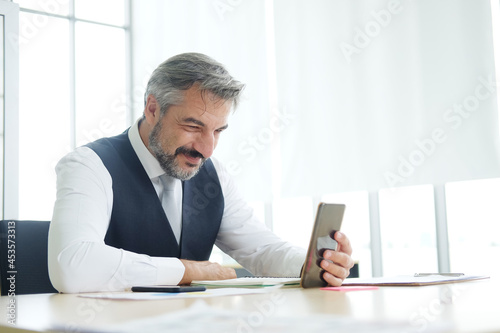 Smart Caucasian senior manager and businessman in black suit relaxing in social media online and talking, calling with businesspeople on smartphone at workspace in office