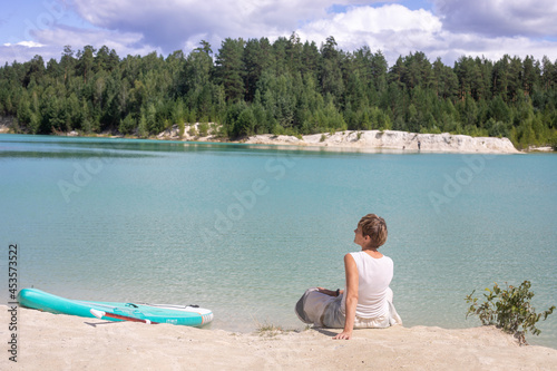 A girl sits on white sand on the shore of a pond with azure-turquoise water. there is a glan board next to it. Vacation and travel in summer