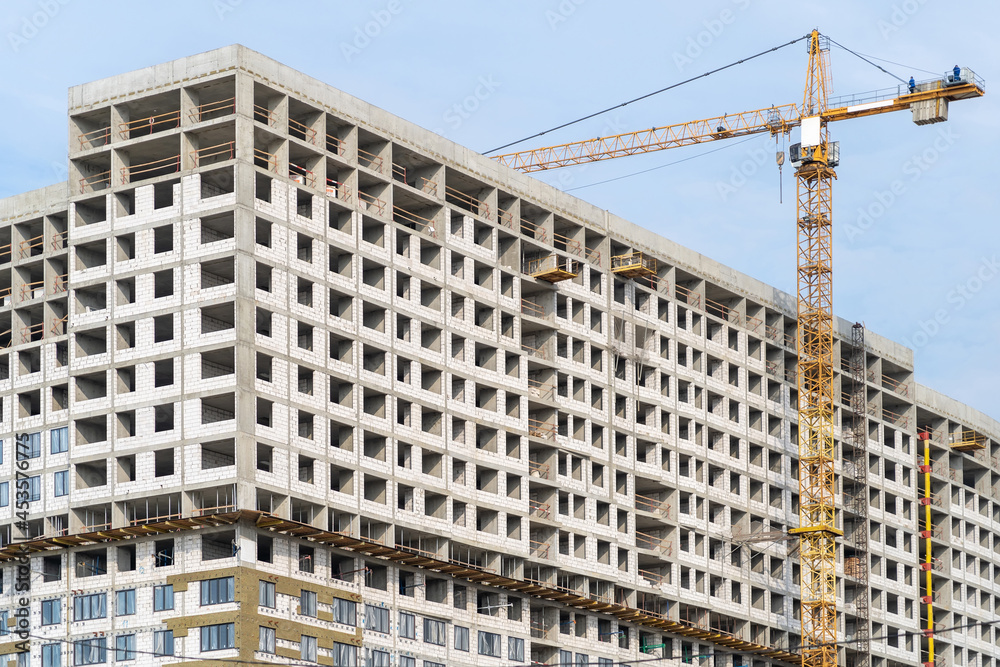 Yellow construction tower crane and unfinished residential building against grey sky. Housing construction, apartment block with scaffolding Yellow construction tower crane and unfinished residential building against grey sky. Housing construction, apartment block with scaffolding