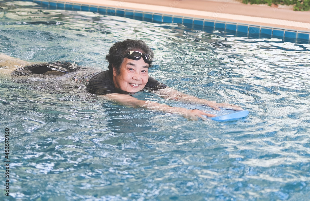 happy and healthy Asian senior woman swimming with kickboard in a swimming pool. Smiling old woman swimming with inflatable board in swimming pool.  Elderly active lifestyle.