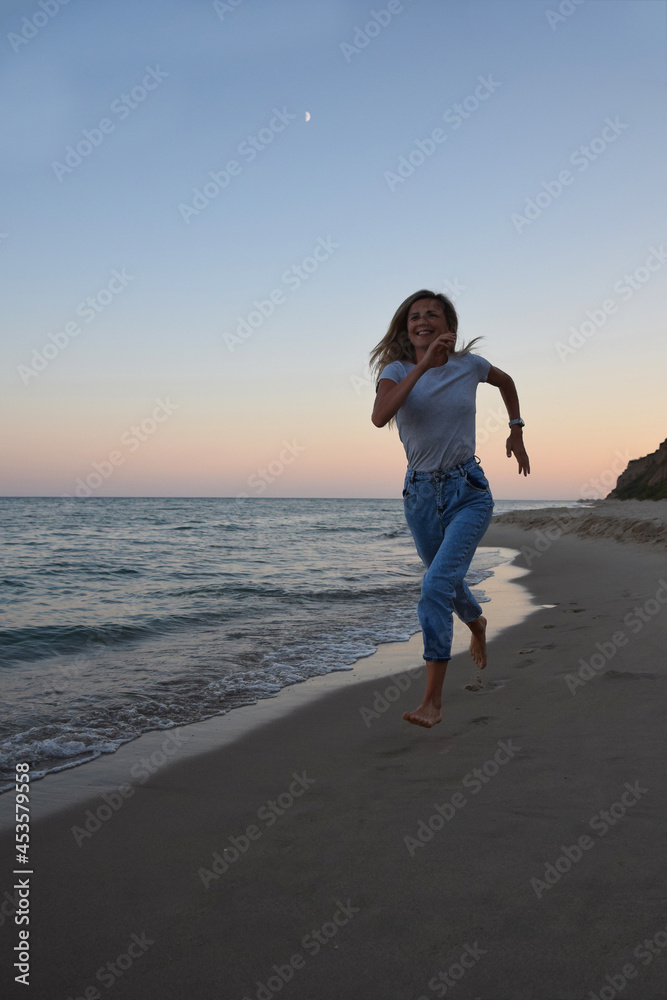 A blonde girl in jeans and a T-shirt is running along the shore of the evening sea