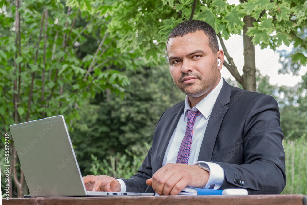 Serious young man in business suit and tie works on open veranda. Business portrait of leading field manager. Busy man with short hair. Middle eastern man organized a mobile office in the park Serious young man in business suit and tie works on open veranda. Business portrait of leading field manager. Busy man with short hair. Middle eastern man organized a mobile office in the park