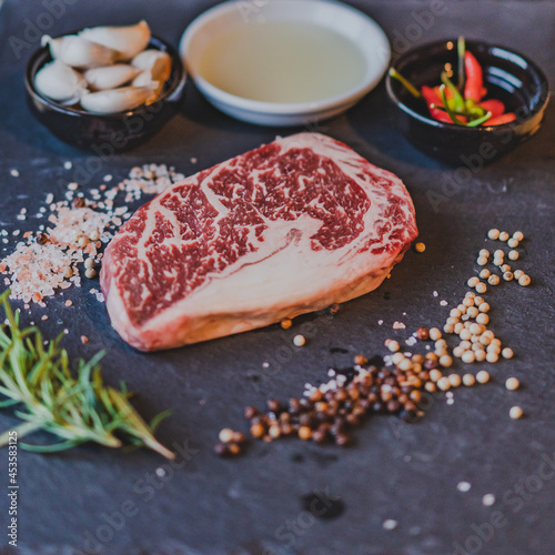 Fresh raw rib eye dry aged steak on stone slate Board with salt, pepper, olive oil,garlic and rosemary.