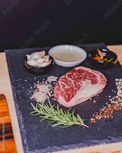 Fresh raw rib eye dry aged steak on stone slate Board with salt, pepper, olive oil,garlic and rosemary.