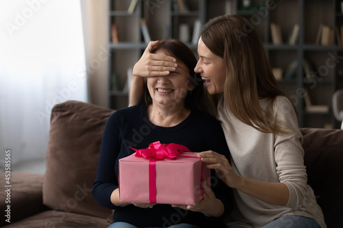 Happy millennial beautiful woman covering eyes of happy middle aged elderly mother, presenting wrapped surprise gift, congratulating with international woman's day or celebrating happy birthday.
