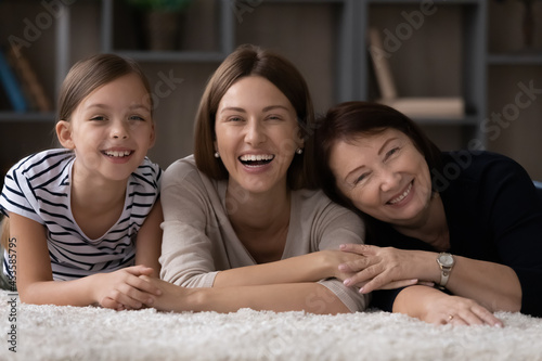 Portrait of happy adorable small kid girl lying on floor carpet with affectionate loving mother and laughing older mature grandmother  enjoying spending carefree weekend leisure time together at home.