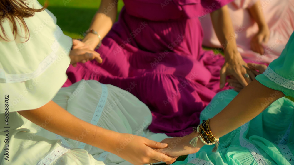 Obraz premium European girls friends in dresses sitting in a circle holding each other's hands in nature with green grass on the background