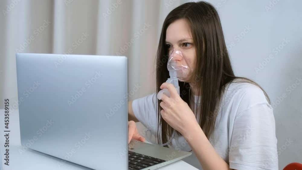Sick young woman is using nebulizer and inhaler for treatment at home and working on laptop. She is breathing in mask. Fibrosis, covid-19, asthma, copd. Treatment, cure and procedure, therapy. Sick young woman is using nebulizer and inhaler for treatment at home and working on laptop. She is breathing in mask. Fibrosis, covid-19, asthma, copd. Treatment, cure and procedure, therapy.