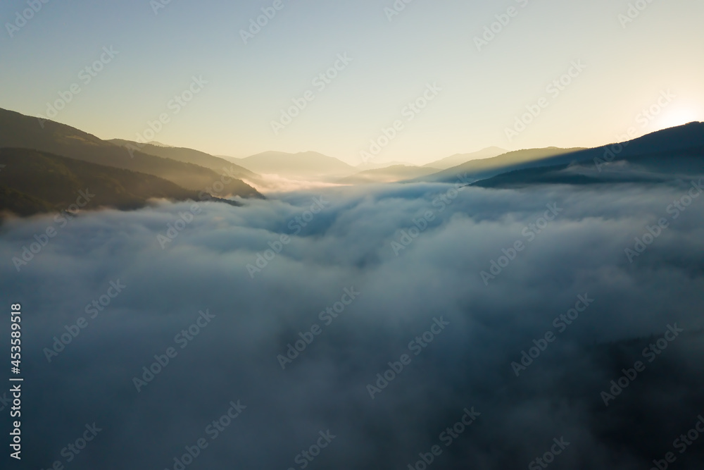 Aerial view of vibrant sunset over white dense foggy clouds with distant dark silhouettes of mountains on horizon.
