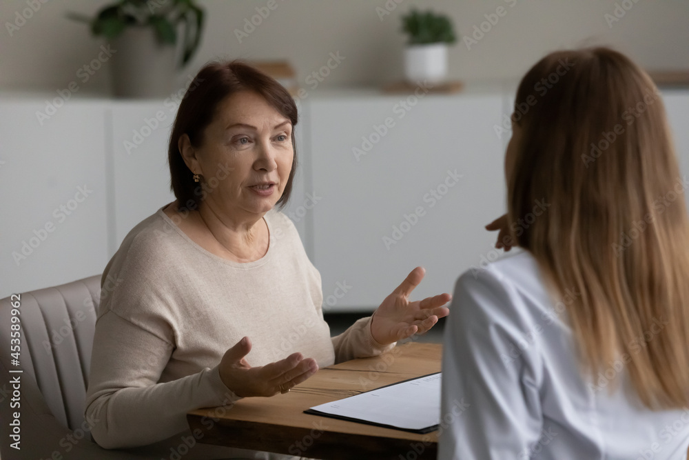 Pleasant beautiful middle aged older retired woman telling health complaints or discussing healthcare issues with young female general practitioner doctor physician at checkup meeting in clinic. Pleasant beautiful middle aged older retired woman telling health complaints or discussing healthcare issues with young female general practitioner doctor physician at checkup meeting in clinic.