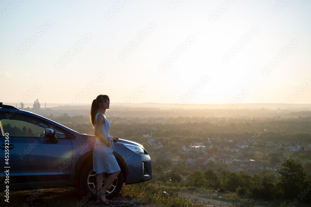Fototapeta premium Happy young woman driver in blue dress leaning on her car enjoying warm summer day. Travelling and vacation concept.
