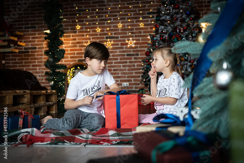 A cute siblings brother and sister in pajamas telling secrets next to a Christmas tree in a decorated room . Children and New Year