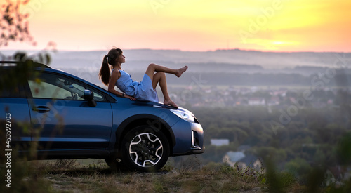 Yong happy female driver resting on hood of her car enjoying sunset view of summer nature. Travel destinations and recreation concept.