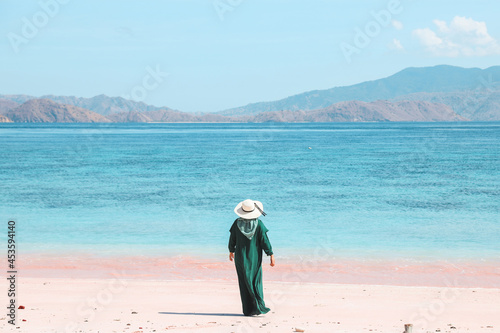 Woman tourist in green dress and summer hat standing on pink sandy beach enjoying seascape and hills at Labuan Bajo