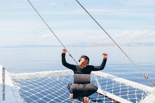 Asian man in wetsuit and headphone sitting on catamaran net while listen to songs in a laptop on vacation at Labuan Bajo