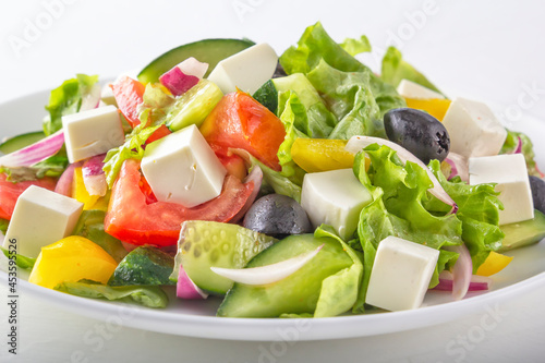 Greek salad of fresh cucumber, tomatoes, bell peppers, lettuce, red onions, feta cheese and olives with olive oil in a white plate on a light background. Healthy eating. Close up
