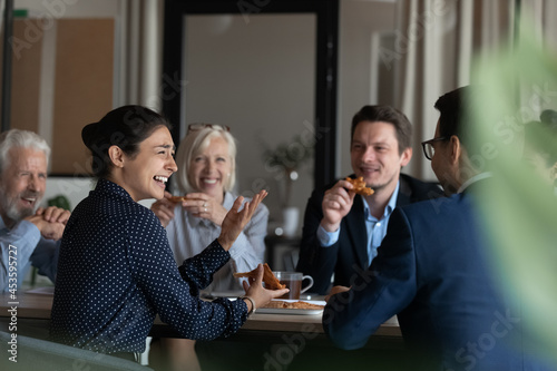 Excited diverse employees eating pizza during break in office together, happy Indian businesswoman laughing at funny joke, talking chatting with colleagues, having fun, sharing corporate lunch
