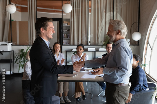 Smiling executive shaking mature business partner hand after group negotiations, colleagues applauding, standing in boardroom, happy team leader congratulating new employee with promotion or new job