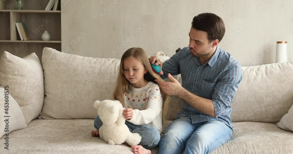 Cute little 9s girl sit on sofa play fluffy bear while young single daddy combing her long hair with modern hairbrush, making hairdo. Family routine, take care of daughter, happy fatherhood concept Cute little 9s girl sit on sofa play fluffy bear while young single daddy combing her long hair with modern hairbrush, making hairdo. Family routine, take care of daughter, happy fatherhood concept