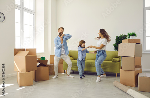 Happy young family having fun in their new home on moving day. Cheerful excited mum, dad and child playing and dancing in room with sofa and unpacked boxes. Real estate, mortgage, buying house concept