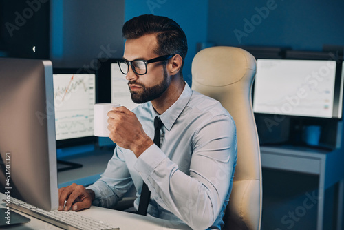 Confident young man drinking coffee and working on computer while staying late in the office